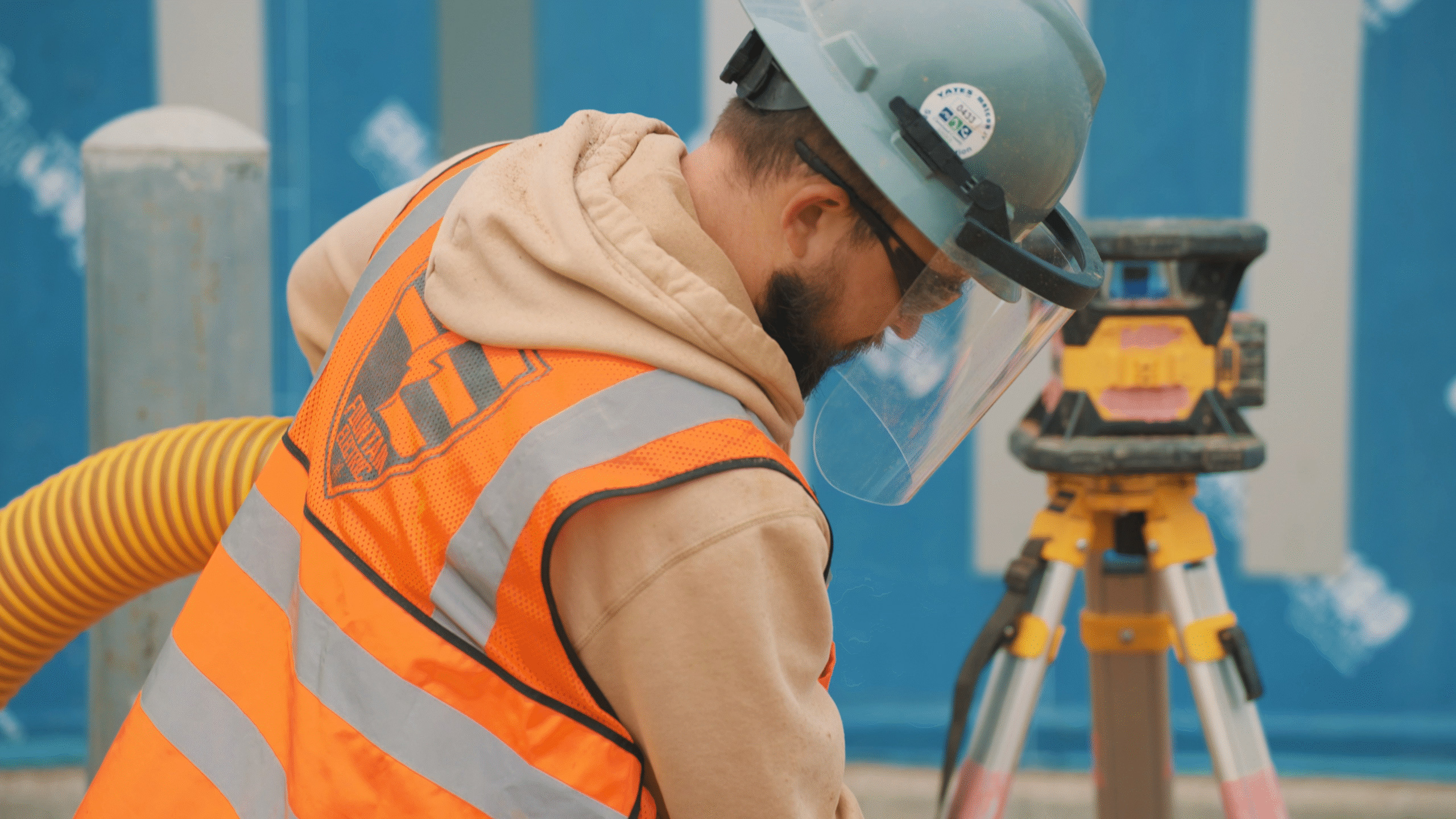 Construction worker wearing hard hat and protective face shield standing near a yellow survey tripod.